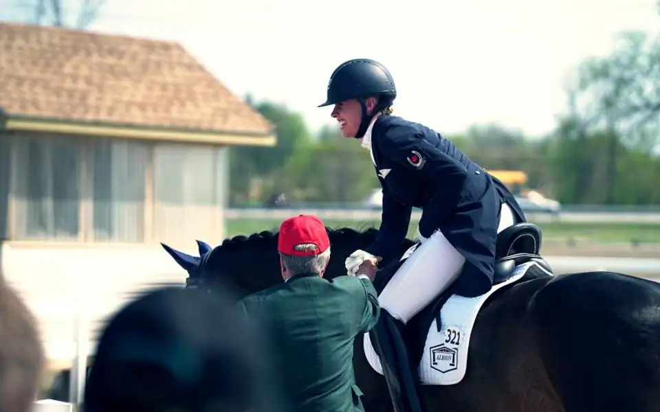 A beginner rider in a black helmet sits on a calm bay horse at a walk in an outdoor sand arena while an instructor stands beside them holding a lead line