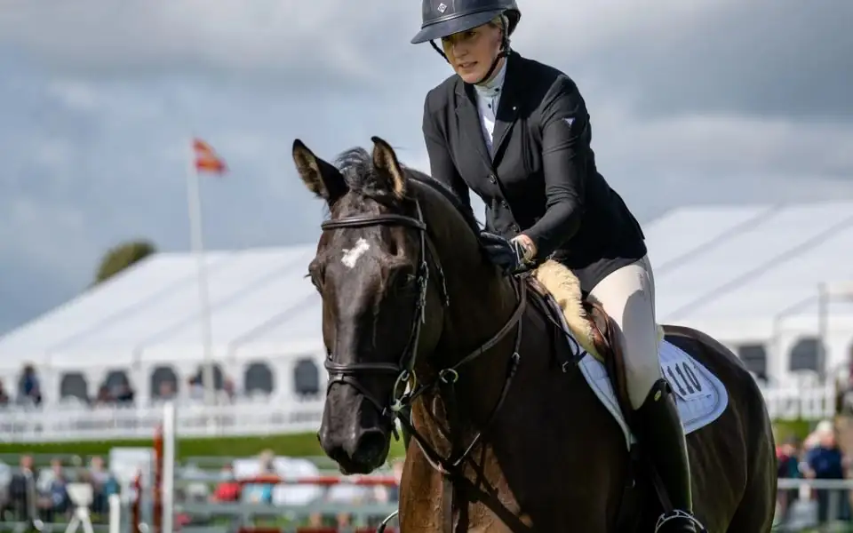 Close-up of a rider fastening the chin harness of a black equestrian helmet while standing beside a saddled grey horse in a barn aisle